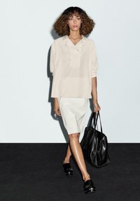 Lichtbeige linnen shirt met knoopsluiting, gecombineerd met een witte kokerrok en een zwarte leren handtas. Zwarte slippers maken de look compleet.