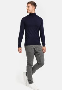 Navy turtleneck sweater in een slim fit, gecombineerd met lichtgrijze slim-fit broek en witte sneakers, staande op een effen achtergrond.