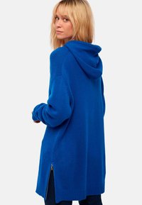 Blaue gestrickte Kapuzenjacke mit lockerer Passform, seitlichen Reißverschlussbelüftungen und gerippten Bündchen. Weicher Stoff und mittellanges Design.