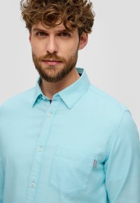 Hellblaues Hemd mit knopffreiem Kragen, aus strukturiertem Stoff, mit einer Brusttasche. Verfügt über dezentes Branding auf der Tasche.