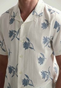 Camicia a manica corta con bottoni in tessuto crema con motivi floreali blu. Presenta un colletto da campo e una superficie leggermente testurizzata.
