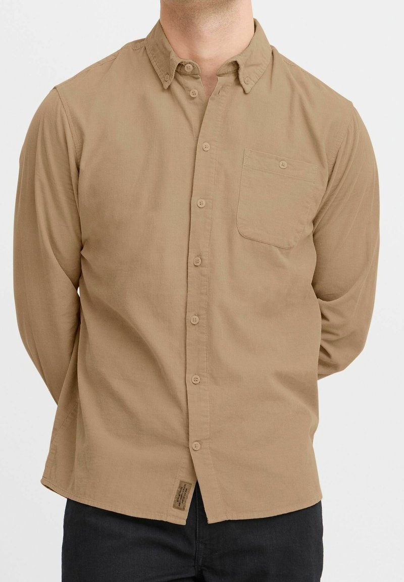 Camisa de manga larga abotonada en tejido marrón claro con un bolsillo en el pecho, una textura sutil y un dobladillo curvado, combinada con pantalones oscuros.