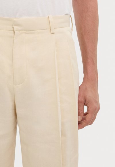 Beige linen trousers feature a flat front, pleats, side pockets, and a waistband with a loop, displayed alongside a partially visible hand.