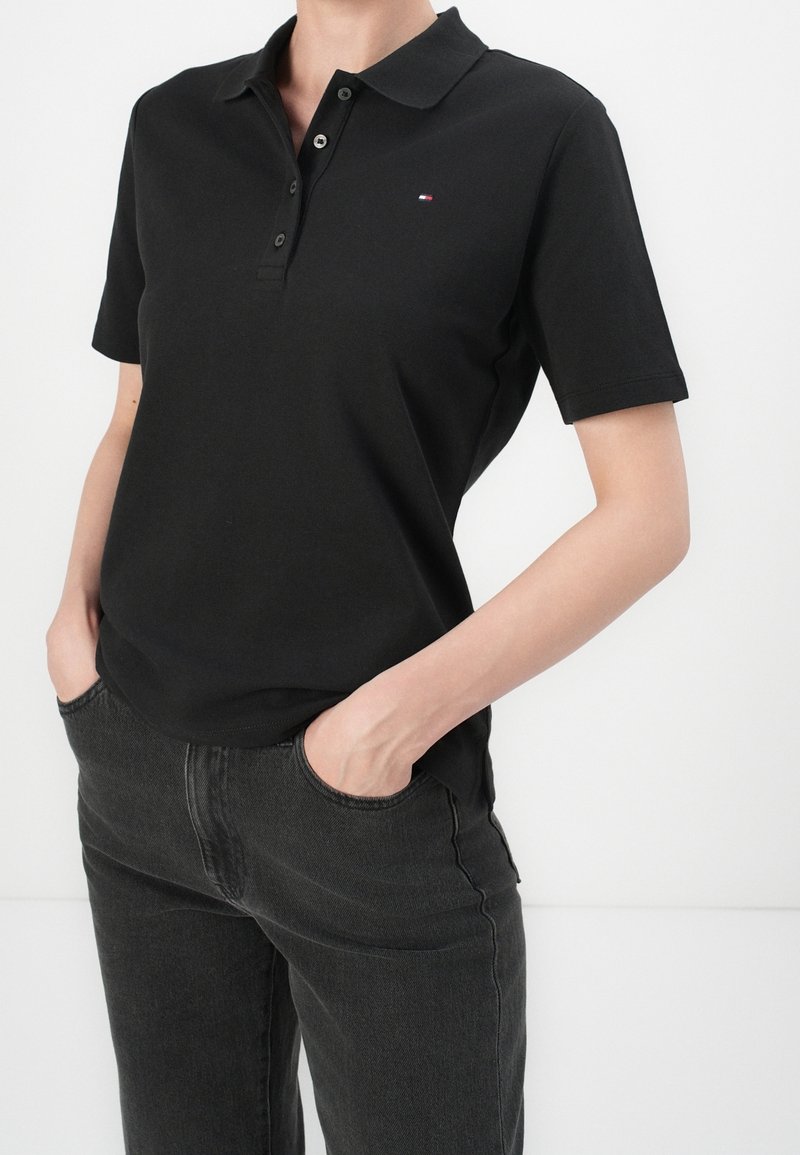 Personne portant un polo noir à manches courtes avec des boutons et un petit logo, associé à un jean foncé, les mains dans les poches devant un fond uni.
