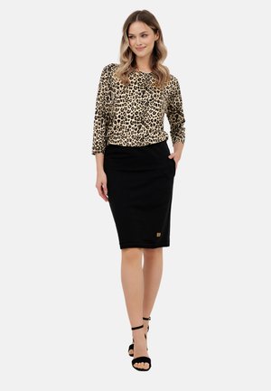 Kobieta w bluzce z długim rękawem w panterkę, czarnej spódnicy do kolan z kieszeniami oraz czarnych szpilkach z paskiem wokół kostki, stojąca na tle białego tła.