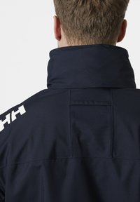 Marineblauwe jas met een hoge kraag, voorzien van een gladde stoftextuur en een wit logo-detail op de linkerschouder.