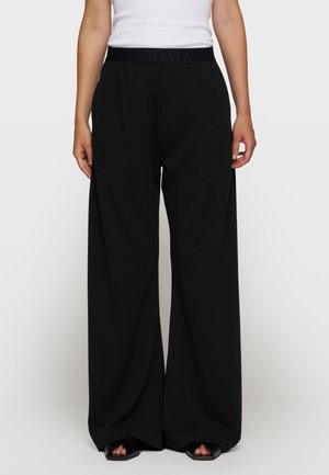 Femme portant un pantalon noir à jambes larges et des chaussures noires à bout ouvert, debout devant un fond blanc uni.