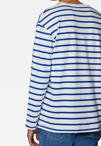 Eine Person trägt ein weißes Langarmshirt mit blauen horizontalen Streifen und blaue Jeans, von hinten vor einem schlichten Hintergrund gezeigt.