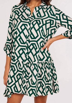 Woman wearing a loose, button-up dress with green geometric patterns on a white background, hand on hip, three-quarter sleeves.