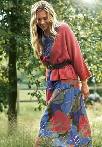 Lange bloemenjurk in blauw met rode en groene patronen, gelaagd onder een geribde roze cardigan, getailleerd met een zwarte riem.