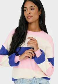 Femme aux longs cheveux noirs portant un pull rayé rose, blanc et bleu et un jean bleu clair, posant avec les mains près de la poitrine.