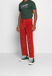 Červené kordové kalhoty rovného střihu, spárované s tmavozeleným tričkem krátkým rukávem s bílým logem a tmavozelenými vysokými teniskami.