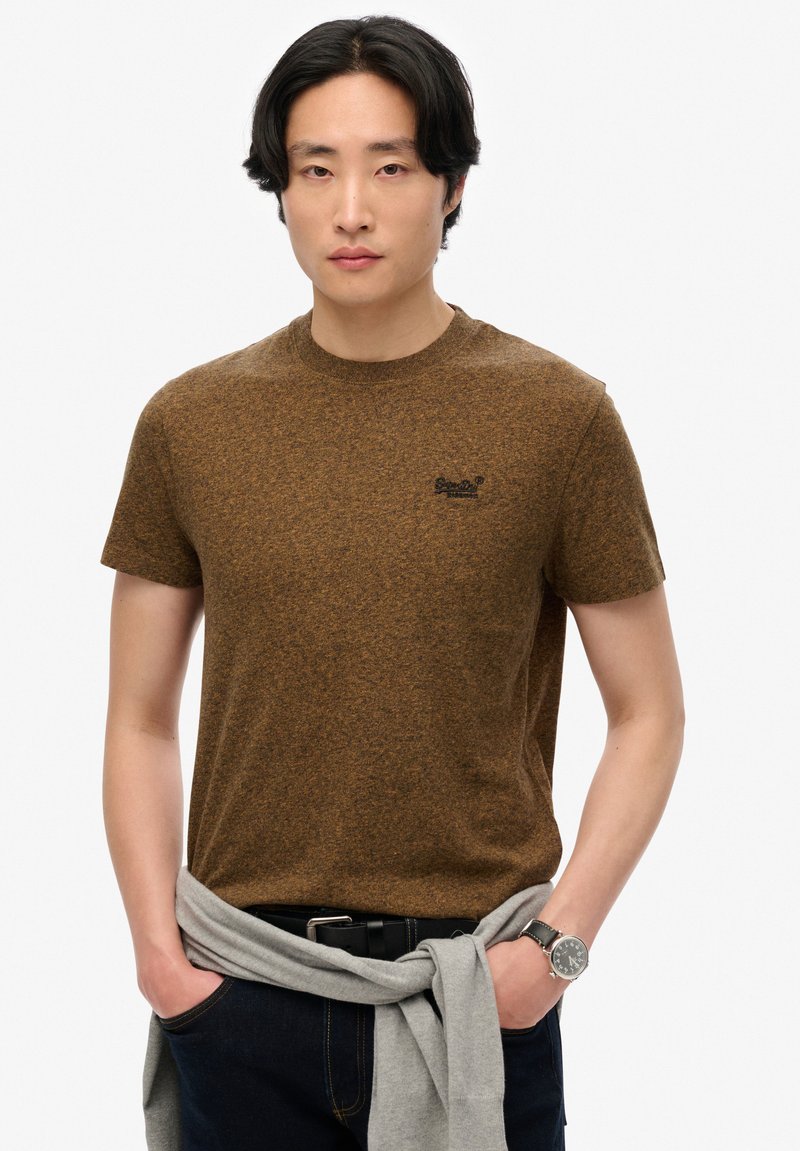 Camiseta marrón jaspeada de manga corta con escote redondo y logo negro en el pecho, combinada con unos jeans oscuros y un suéter gris atado a la cintura.
