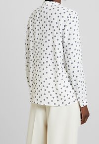 White polka dot shirt with gray dots, long sleeves, and a rounded hem. Made of smooth fabric, featuring button cuffs and a classic collar.
