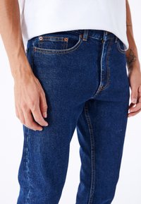 Jeans de mezclilla de color azul oscuro con fit ajustado. Incluyen costuras de cobre, diseño de cinco bolsillos y cierre de botones. La textura es suave y ligeramente áspera.