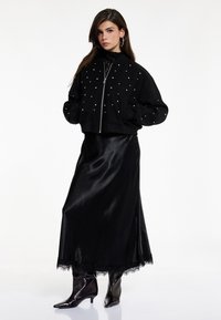 Schwarze, kurze Bomberjacke mit silbernen Nieten, getragen über einem schwarzen Satin-Maxikleid mit Spitzenbesatz, kombiniert mit schwarzen spitzen Stiefeletten.