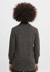 Person med lockigt hår iförd en mörkgrå tröja med långärmar, stående med ryggen mot en enkel ljus bakgrund.