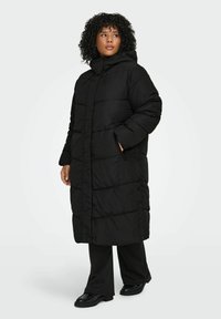 Melns pūkains mētelis ar kapuci, ar quiltētu rakstu, priekšpuses rāvējslēdzēju un aizdares pogām. Modelis valkā melnas bikses un kurpes.