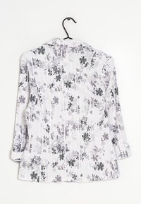 Blouse à motif floral en tissu blanc avec des accents gris. Présente un col et des manches longues avec des poignets à boutons.
