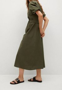 Olivengrünes, knielanges Kleid mit puffärmeln und tailliertem Bund. Getragen mit schwarzen flachen Sandalen mit einer Schnallenverzierung.