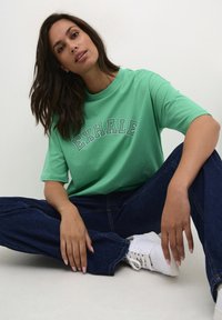 Groene katoenen t-shirt met de tekst "EXHALE" in zwart omrandde letters, gecombineerd met donkerblauwe wijde jeans en witte sneakers, zittende pose.