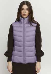 Dikke vest in lichtpaars, met hoge kraag en rits aan de voorkant. Gecombineerd met een donkerbruin gebreid sweater, met gestructureerde mouwen.