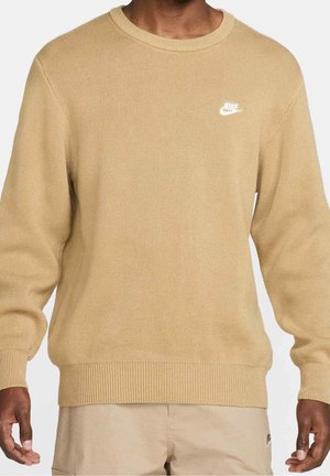 Uomo che indossa un maglione in maglia beige di Nike con colletto, polsini e orlo a coste, abbinato a pantaloni beige su uno sfondo neutro.