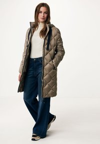 Gequilted beige lange pufferjas met een capuchon, zwarte trekkoorden, zijzakken, gedragen over een witte top en blauwe uitlopende jeans. Geweven stof.