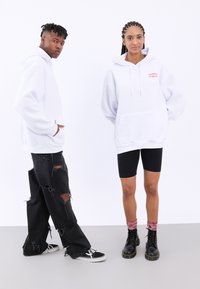 Felpe oversized bianche con tasca frontale, jeans neri strappati e stivaletti neri; il modello a sinistra ha un tono di pelle chiaro, mentre il modello a destra ha un tono di pelle più scuro.