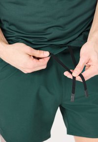 Grüne Sportshorts mit strukturierter Oberfläche. Hände justieren die schwarzen Kordeln mit Gummispitzen am Bund.