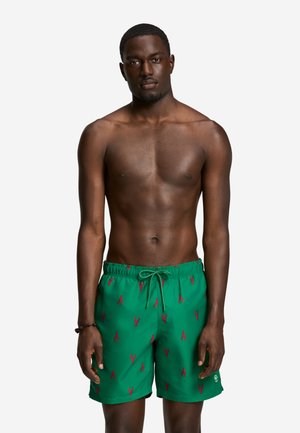 Jeune homme à la peau foncée portant un short de bain vert avec des motifs de homards rouges, debout devant un fond blanc.