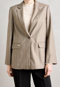 Hnědý blazer s proužky a strukturovanými rameny, přední zapínání na knoflík, dvě klopové kapsy a zlaté iniciály "M.M." na levé kapse.