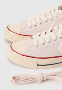 Sneaker in tela rosa con punta in gomma, suola beige, dettagli con striscia rossa e lacci color crema. Include un ulteriore laccio rosa annodato nelle vicinanze.