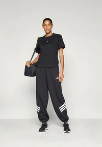 Ženska stoji oblečena v črno majico Adidas, črne trenirke Adidas z belimi stranicami, črne superge in nosi črno torbo čez ramo.