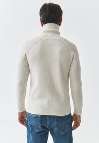Pullover cu tricotaj în ramă de culoare crem, cu guler înalt, având mâneci raglan și o margine ajustată, asociat cu blugi din denim albastru.