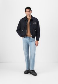 Marineblauer Bomberjacke mit Druckknöpfen, zwei Fronttaschen, einem Aufnäher auf der Brust, kombiniert mit einem beigen Pullover und hellblauen Jeans.
