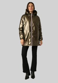 Doudoune brillante de couleur olive, avec capuche, fermeture éclair à l'avant et poches latérales. Portée sur un pantalon noir évasé et des bottines.