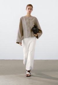Beige linnen top met een boxy pasvorm, vijf knoopdetails en brede mouwen, gecombineerd met losse witte broek en een zwarte, geprinte clutch.