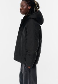 Giacca nera con cappuccio, chiusura con zip, texture trapuntata e tasche laterali. Indossata con pantaloni della tuta grigio scuro per un look casual.