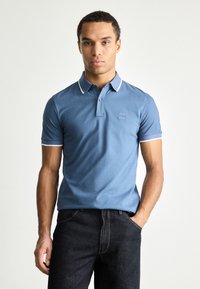 Polo shirt bleu avec col, manches courtes et accents de bord blanc. Présente une petite patch logo sur la poitrine. Texture douce, design ajusté.