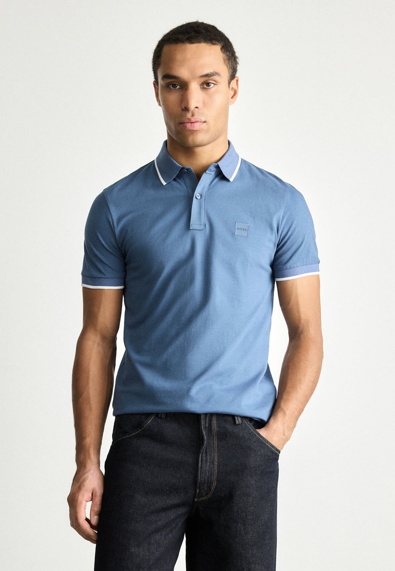 Camisa polo azul com gola, mangas curtas e detalhes em branco. Apresenta um pequeno patch com o logótipo no peito. Textura suave, design ajustado.