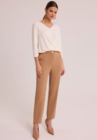 Kremowa bluzka z dekoltem w kształcie V i rękawami o długości trzech ćwierć, w parze z beżowymi spodniami z wysokim stanem. Klasyczny krój o gładkiej fakturze i zwężającym się fasonie nogawek.