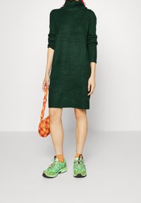 Abito a collo alto, lavorato a maglia, di un verde scuro con maniche lunghe. Abbinato a sneaker verde brillante e una borsa a quadri arancione. Texture morbida e vestibilità ampia.