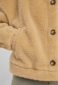 Giacca in pile soffice e di colore beige, con una superficie testurizzata, dotata di grandi bottoni e tasche laterali. L'orlo e i polsini sono morbidi e aderenti.