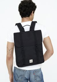 Svart tygryggsäck med justerbara remmar, viklockstängning och metallisk logodetalj. Har en minimalisk design och en strukturerad form.