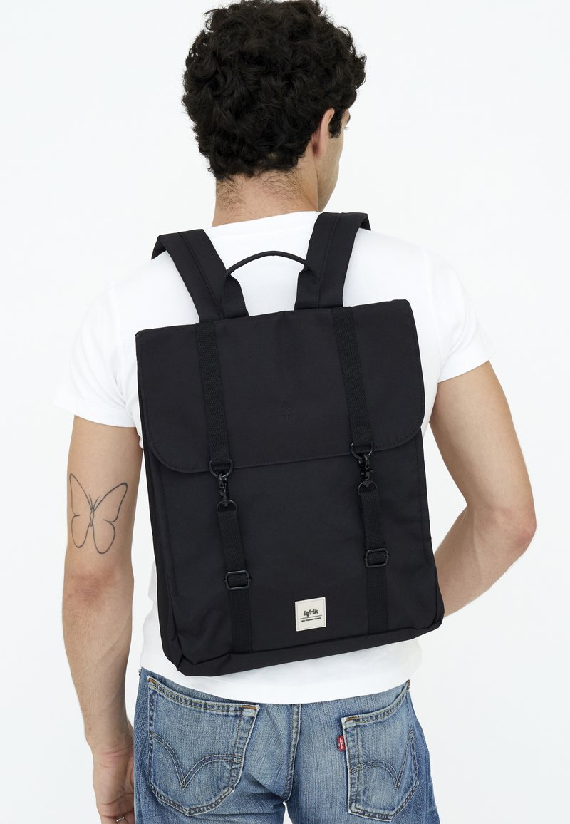 Svart tygryggsäck med justerbara remmar, viklockstängning och metallisk logodetalj. Har en minimalisk design och en strukturerad form.