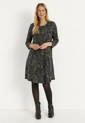 Kvinde stående iført en mørk kjole med paisley-mønster og 3/4-ærmer, sorte strømpebukser og sorte ankelstøvler, hænder løst foldet.