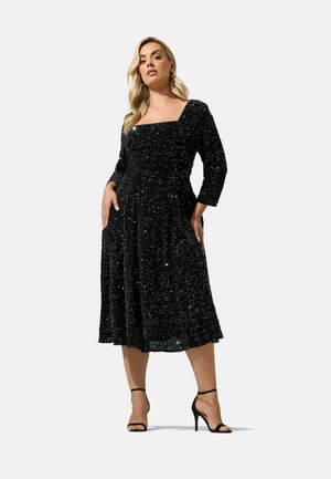Mujer que lleva un vestido midi negro de manga larga con lentejuelas, escote cuadrado y sandalias negras de tacón alto.