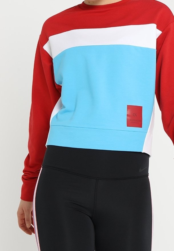 Personne portant un sweat-shirt color-block avec des manches rouges, un devant blanc et bleu, et des leggings noirs avec des bandes latérales.