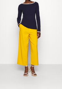 Žena v námořnickém svetru s dlouhým rukávem a bílým lemem, v jasně žlutých širokých kalhotách a bílých sandálech na vysokém podpatku se řemínky.